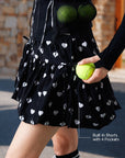 Close-up of Pom Pêche black Bubble Skirt showing built-in shorts with 4 pockets holding tennis balls – Pom Pêche ribbon waistband and heart print fabric for court play