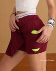 Close-up of Pom Pêche wine red Classic Pleated Skirt with 4 Hidden Pockets callout – model lifting pleated outer skirt to reveal built-in shorts with dual side pockets holding yellow tennis balls, Pom Pêche wristband
