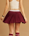 Back view of Pom Pêche Classic Pleated Tennis Skirt in wine red – fine knife pleats fanning out from Pom Pêche white logo waistband with pink and black stripe, styled with cream racerback tank and wine red Pom Pêche knee socks