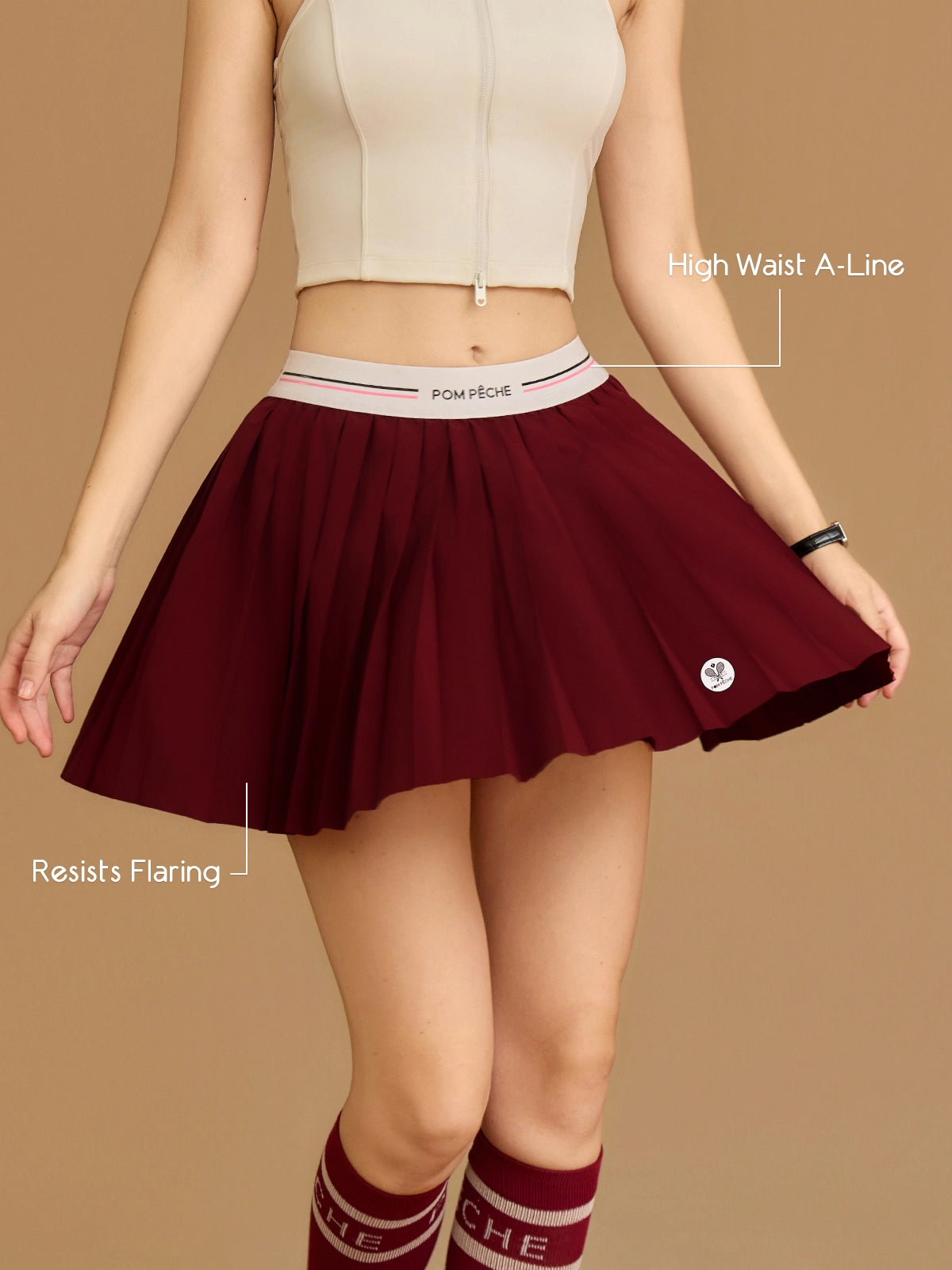 Pom Pêche wine red Classic Pleated Tennis Skirt feature callout – High Waist A-Line silhouette and Resists Flaring technology annotated, model holding out pleated hem showing structured A-line shape