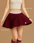 Pom Pêche wine red Classic Pleated Tennis Skirt feature callout – High Waist A-Line silhouette and Resists Flaring technology annotated, model holding out pleated hem showing structured A-line shape