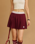 Pom Pêche Classic Pleated Tennis Skirt in wine red – fine knife pleats with Pom Pêche white logo waistband with pink and black stripe, racket badge emblem, styled with cream zip crop top and Pom Pêche wine red knee socks