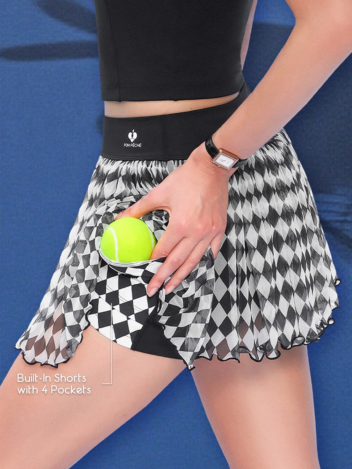 Close-up of Pom Pêche black Diamond Mesh Skirt with Built-In Shorts with 4 Pockets callout – model lifting harlequin diamond mesh skirt to reveal black built-in shorts pocket holding yellow tennis ball, Pom Pêche script waistband