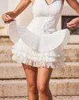 Pom Pêche Spin™ LaRose white pleated tiered skirt – model holding out fan-shaped fine pleated outer layer revealing three-tier mesh underlayers, styled with zip halter top on Paris stone steps