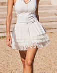 Full body view of Pom Pêche Spin™ LaRose white pleated tiered skirt – high-waisted sculpting waistband with metallic Pom Pêche logo, styled with zip halter top on Paris stone steps