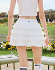 Back view of Pom Pêche Spin™ Mille white tiered skirt – Pom Pêche crown logo on waistband, 4-layer tulle ruffle hem, styled with white crop tank and striped tennis socks in Paris garden