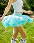 Side view of Pom Pêche mint green Spin™ Petal Tiered Skirt in motion – three-layer asymmetric chiffon petal hem flaring during spin, styled with white zip halter top and white bow knee socks in outdoor park
