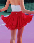 Back view of Pom Pêche wine red Spin™ Petal Tiered Skirt – three-layer chiffon petal hem flaring, high waistband with Pom Pêche heart crystal stud detail, styled with white open-back Poise halter polo top on purple clay court