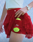 Close-up of Pom Pêche wine red Petal Tiered Skirt with Built-In Shorts with 4 Pockets callout – model lifting chiffon skirt to reveal built-in shorts with dual side pockets each holding a yellow tennis ball, Pom Pêche crown logo print