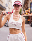 Close-up of Pom Pêche white Poise™ Mesh Back Tank with feature callouts – Deep U Neck and Built-In Removable Bra annotated, Pom Pêche Sports Club Paris burgundy script print, model holding tennis racket on Paris street