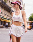 Full body view of Pom Pêche Poise™ Mesh Back Tank in white – Deep U neckline, Pom Pêche Sports Club Paris burgundy script print, styled with white Sports Club shorts, pink Pom Pêche visor and pearl necklace walking on Paris street with tennis racket