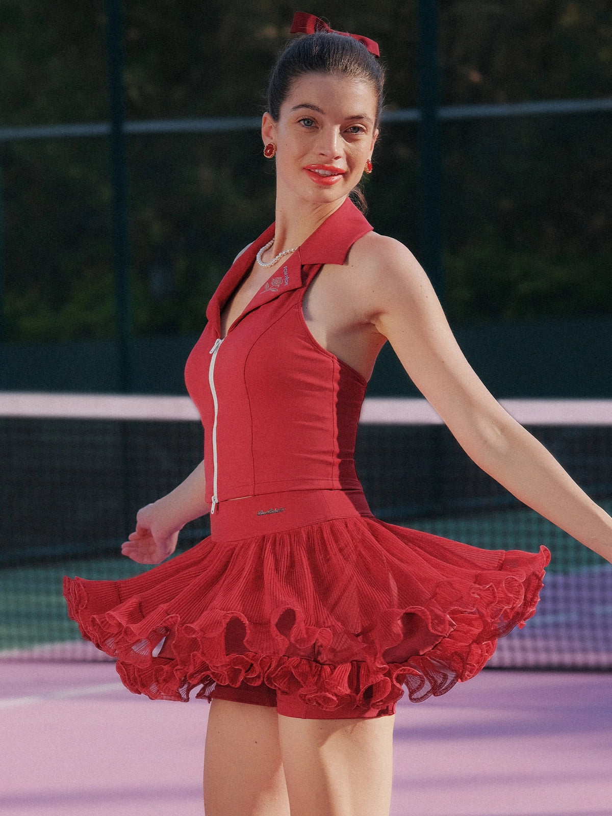 Side view of Pom Pêche Paris red Poise™ Rose Halter Polo Tank – open-back halter silhouette with dual-zipper open at waist, styled with wine red Rose Ruffle Skirt flaring in motion on outdoor tennis court
