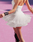 Back view of Pom Pêche white Rose Ruffle Skirt in motion – V-shaped sculpting back waistband with Pom Pêche script logo, voluminous pleated mesh ruffle hem flaring, styled with white halter top and Pom Pêche wristband on pink court