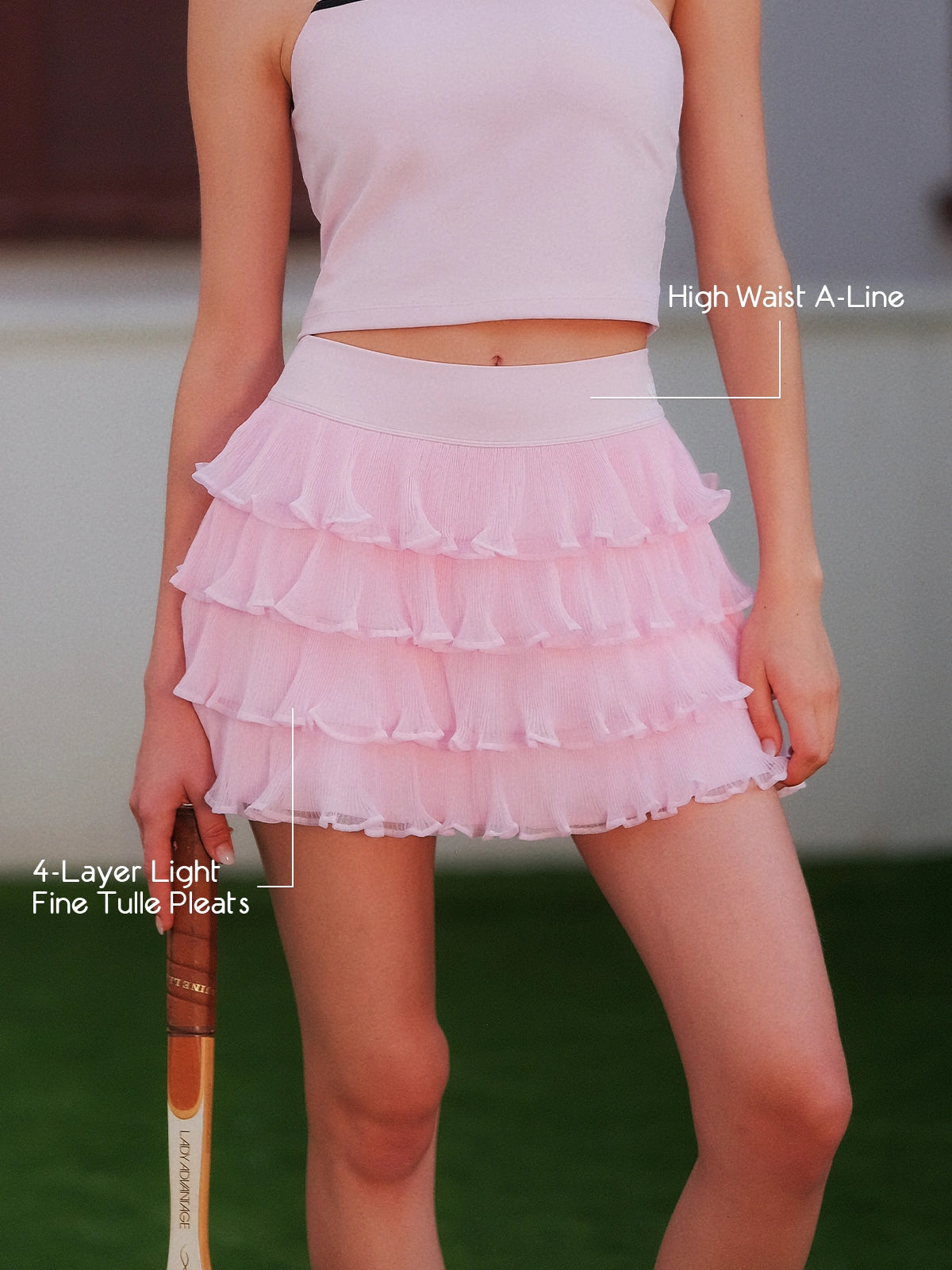 Pom Pêche pink Ruffle Cake Skirt feature callout – high waist A-line silhouette and 4-layer light fine tulle pleats annotated, model holding tennis racket on grass court