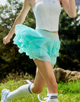 Pom Pêche Spin™ Petal Tiered Tennis Skirt in mint green – three-layer featherlight chiffon petal tiers flaring during run, high-waisted mint band with Pom Pêche logo, styled with white zip halter top and white stripe knee socks on grass