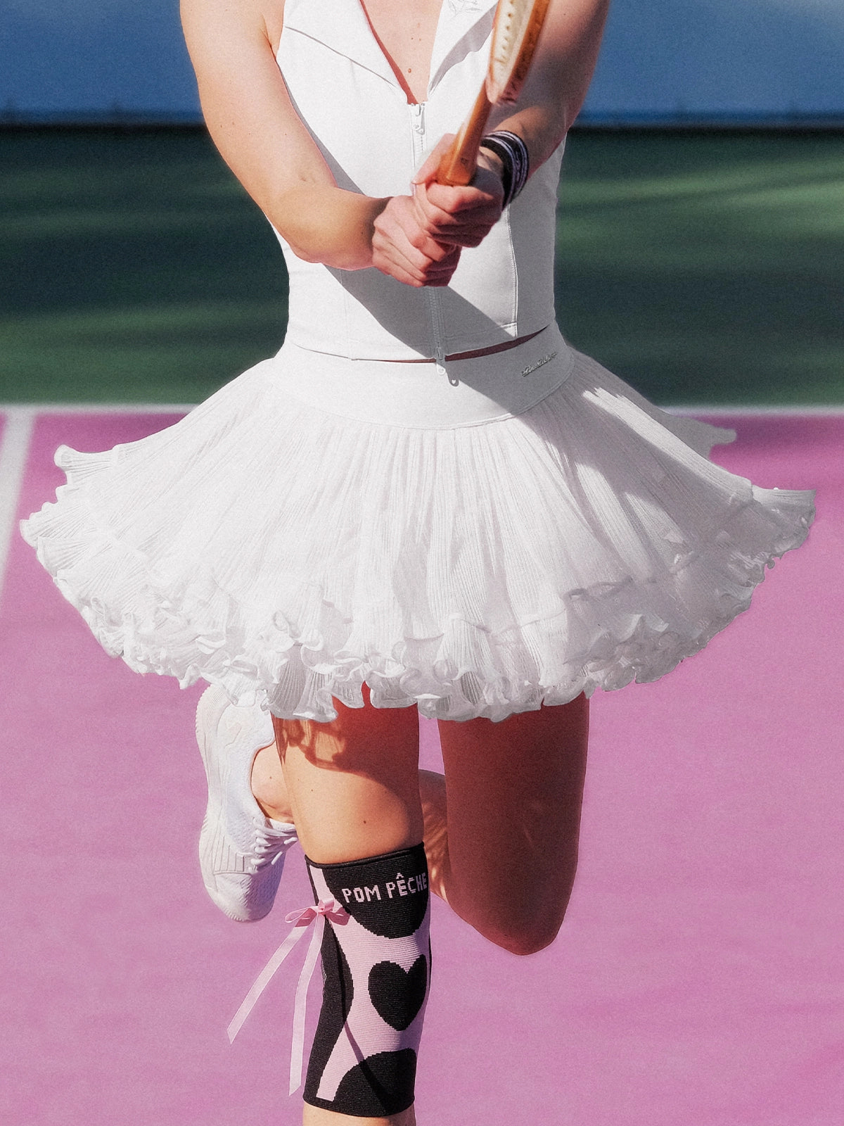 Pom Pêche Spin™ Rose Ruffle Skirt in tennis white – voluminous pleated mesh ruffle hem flaring during tennis swing, styled with white Poise zip halter top and Pom Pêche black heart knee socks on pink clay court