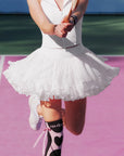 Pom Pêche Spin™ Rose Ruffle Skirt in tennis white – voluminous pleated mesh ruffle hem flaring during tennis swing, styled with white Poise zip halter top and Pom Pêche black heart knee socks on pink clay court