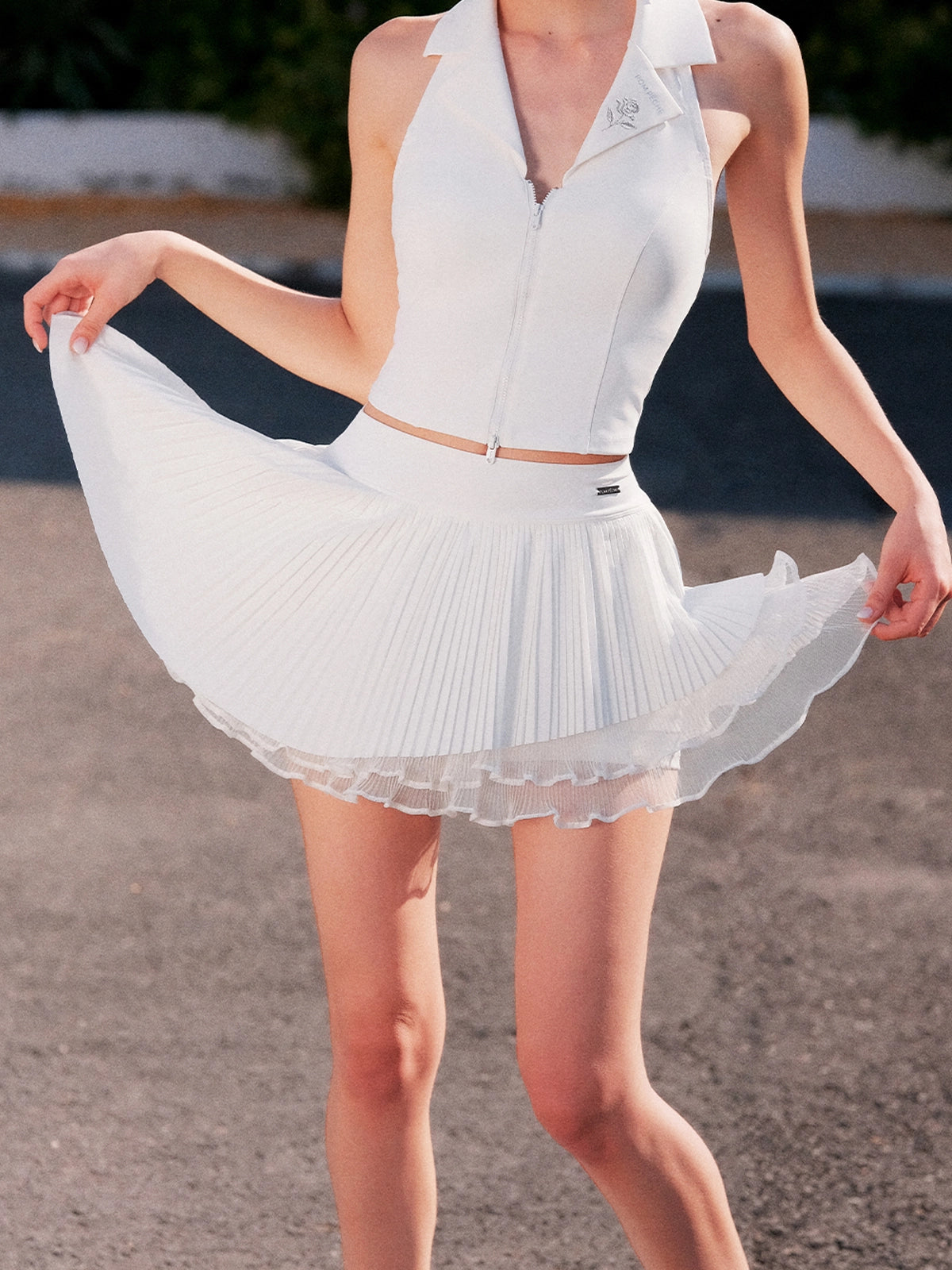 Pom Pêche Spin™ Waltz Layered Tennis Skirt in white – model holding out multi-layer asymmetric pleated skirt showing voluminous A-line silhouette, high-waisted band with Pom Pêche metal logo tag, styled with white Poise polo zip halter top on outdoor court