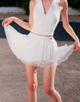 Pom Pêche Spin™ Waltz Layered Tennis Skirt in white – model holding out multi-layer asymmetric pleated skirt showing voluminous A-line silhouette, high-waisted band with Pom Pêche metal logo tag, styled with white Poise polo zip halter top on outdoor court