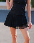 Pom Pêche Spin™ Waltz black pleated layered skirt – front view showing high-waisted silhouette with metallic logo detail and asymmetric tiered hem, styled with black halter tank on court