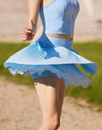 Pom Pêche Spin™ Waltz sky blue layered skirt in motion – multi-layer asymmetric pleated hem flaring during run revealing built-in shorts, styled with matching blue zip halter top on clay court