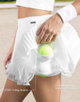Close-up of Pom Pêche white Waltz Layered Skirt with LYCRA® 4-Way Stretch and Four Side Pockets holds 8 balls at once callouts – model lifting pleated skirt to reveal built-in shorts pocket holding yellow tennis ball, Pom Pêche wristband detail