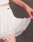 Macro detail of Pom Pêche white Waltz Layered Tennis Skirt showing double-layer construction – outer fine pleated LYCRA® layer over inner semi-sheer mesh ruffle scalloped hem, smooth anti-static 4-way stretch fabric