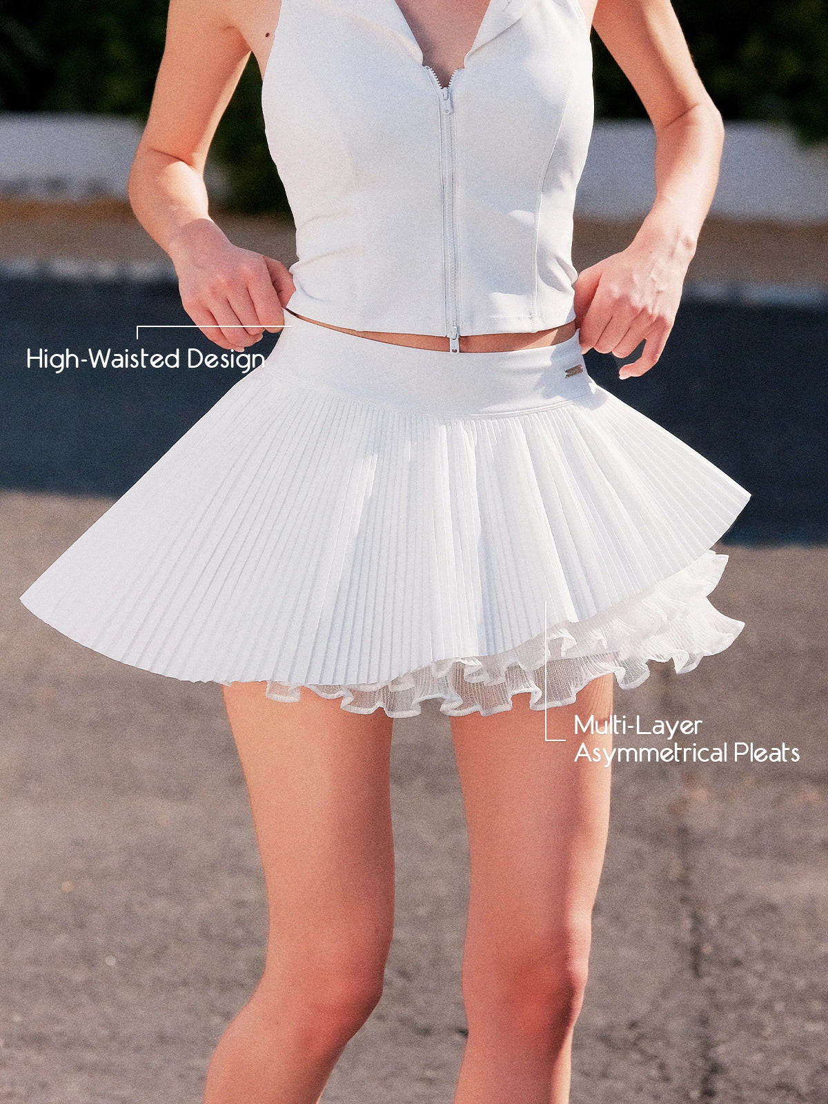 Pom Pêche white Waltz Layered Skirt feature callout – High-Waisted Design and Multi-Layer Asymmetrical Pleats annotated, model pulling waistband showing sculpting high-waist band with Pom Pêche metal tag and double-layer asymmetric pleated hem on outdoor court