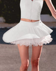 Pom Pêche white Spin™ Waltz Layered Skirt in motion – double-layer asymmetric pleated hem flaring dramatically during spin, high-waisted band with Pom Pêche metal logo tag, styled with white Poise polo zip halter top on outdoor court poolside