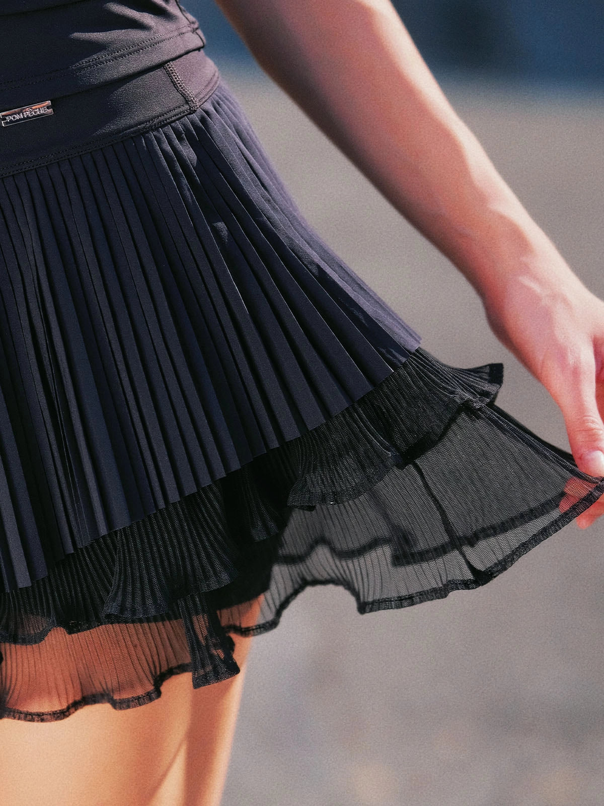 Macro detail of Pom Pêche black Waltz Skirt showing fine pleated LYCRA® outer layer over double-tier sheer mesh ruffle hem – smooth anti-static finish with Pom Pêche logo waistband