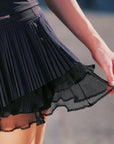Macro detail of Pom Pêche black Waltz Skirt showing fine pleated LYCRA® outer layer over double-tier sheer mesh ruffle hem – smooth anti-static finish with Pom Pêche logo waistband