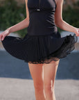 Pom Pêche Spin™ Waltz black layered tennis skirt – model holding out fine pleated outer layer revealing sheer mesh ruffle underlayer, styled with black halter tank on outdoor court