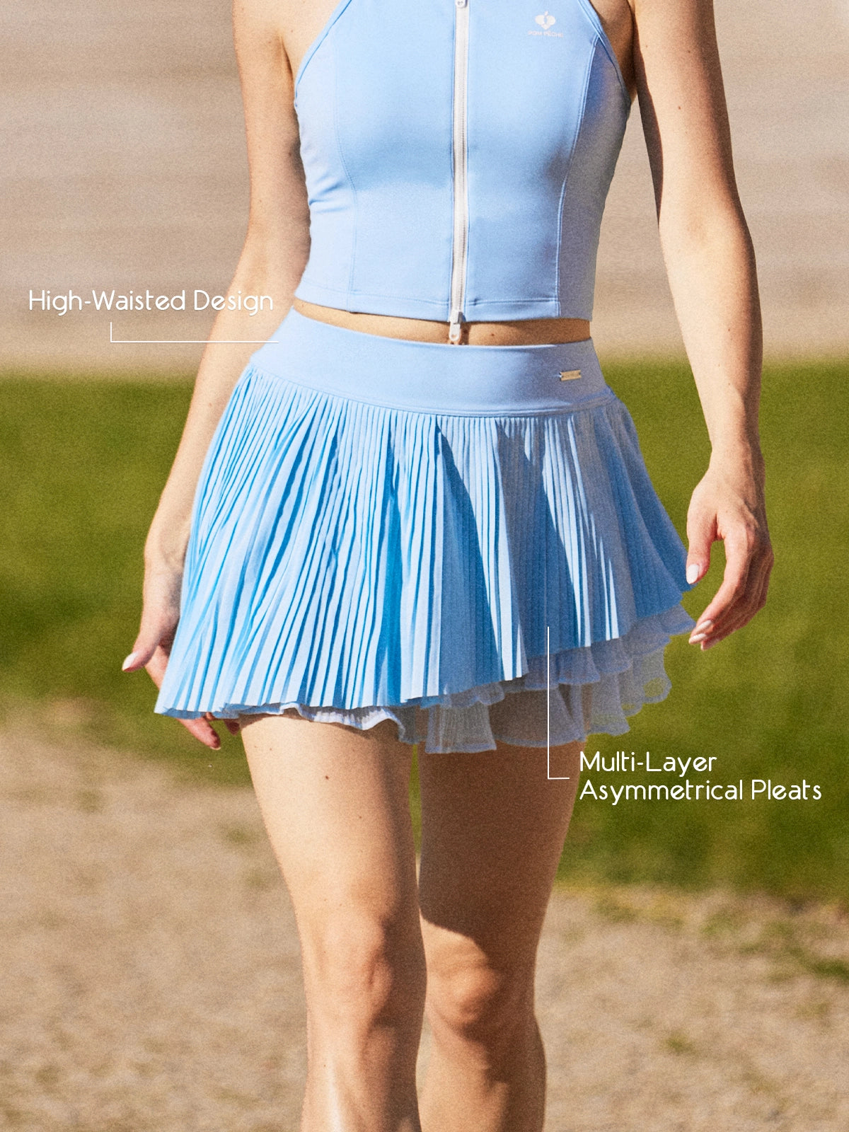 Pom Pêche sky blue Waltz Skirt feature callout – High-Waisted Design for elongating silhouette and Multi-Layer Asymmetrical Pleats for graceful movement annotated, styled with matching blue zip halter top on clay court