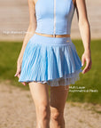 Pom Pêche sky blue Waltz Skirt feature callout – High-Waisted Design for elongating silhouette and Multi-Layer Asymmetrical Pleats for graceful movement annotated, styled with matching blue zip halter top on clay court
