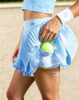 Close-up of Pom Pêche sky blue Waltz tennis skirt showing four side pockets holding tennis balls – built-in shorts with Pom Pêche metallic logo waistband, styled with Pom Pêche wristband