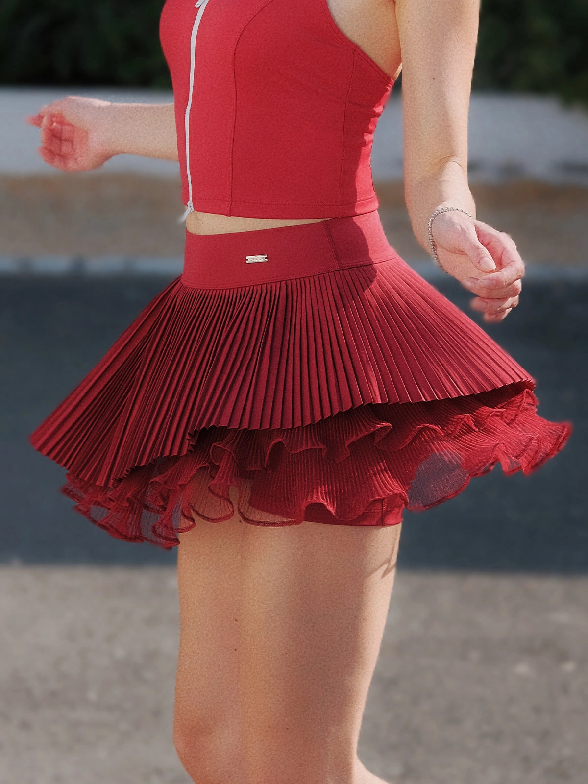 Pom Pêche Spin™ Waltz wine red layered tennis skirt in motion – multi-layer asymmetric pleats flaring with movement, styled with matching zip halter top and striped socks