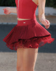 Pom Pêche Spin™ Waltz wine red layered tennis skirt in motion – multi-layer asymmetric pleats flaring with movement, styled with matching zip halter top and striped socks