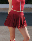 Full body view of Pom Pêche Spin™ Waltz wine red pleated tennis skirt – high-waisted asymmetric silhouette styled with matching zip top and red striped tennis socks on court