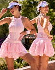 Pom Pêche Zip-Neck Halter Racerback Tank in blush pink – front and back view showing high halter neckline with front zip and blue floral emblem, open racerback with back phone pocket holding pink phone, styled with pink Rose Ruffle Skirt and Pom Pêche visor in Paris garden