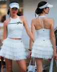 White Bow Halter Tennis Top by Pom Pêche, front lifestyle shot on grass court with contrast black bow and white tennis skirt