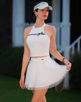 White Bow Halter Tennis Top with matching white tennis skirt, model lifting skirt hem on grass court, Pom Pêche court-to-street look